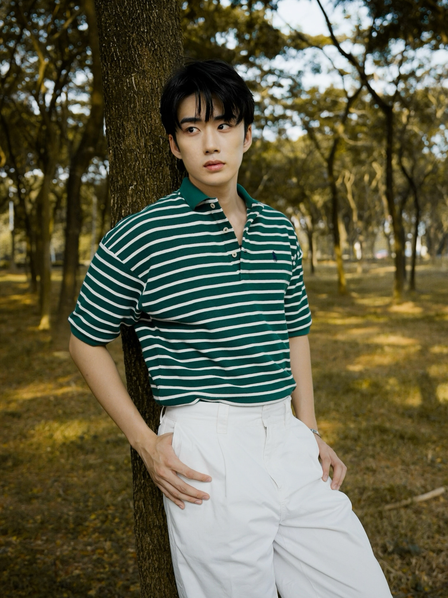 YANG YAN in a green striped polo shirt leaning against a tree in a park
