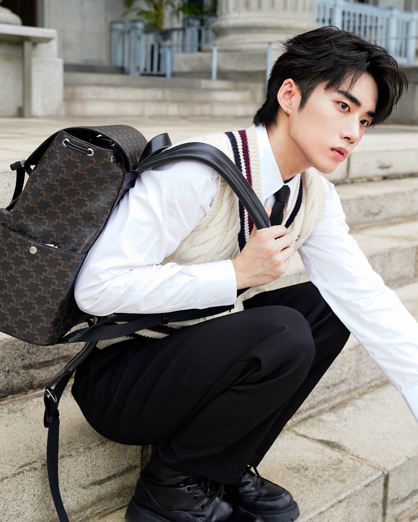 YANG YAN in a knit vest and tie with a monogram backpack on stone steps