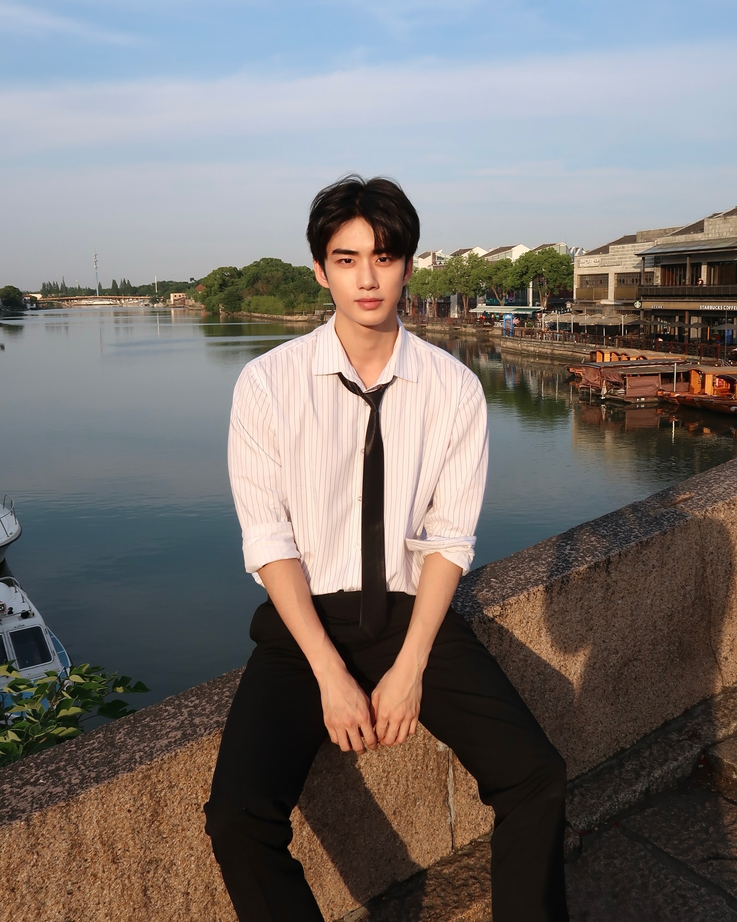 YANG YAN in a striped shirt and loosened tie sitting by a riverside
