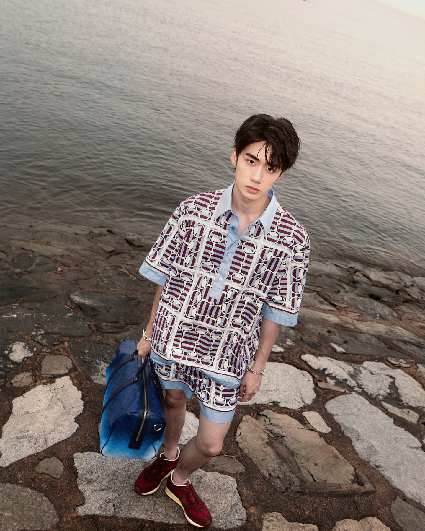 YANG YAN in a patterned short-sleeve shirt with a blue duffel bag on a rocky shore