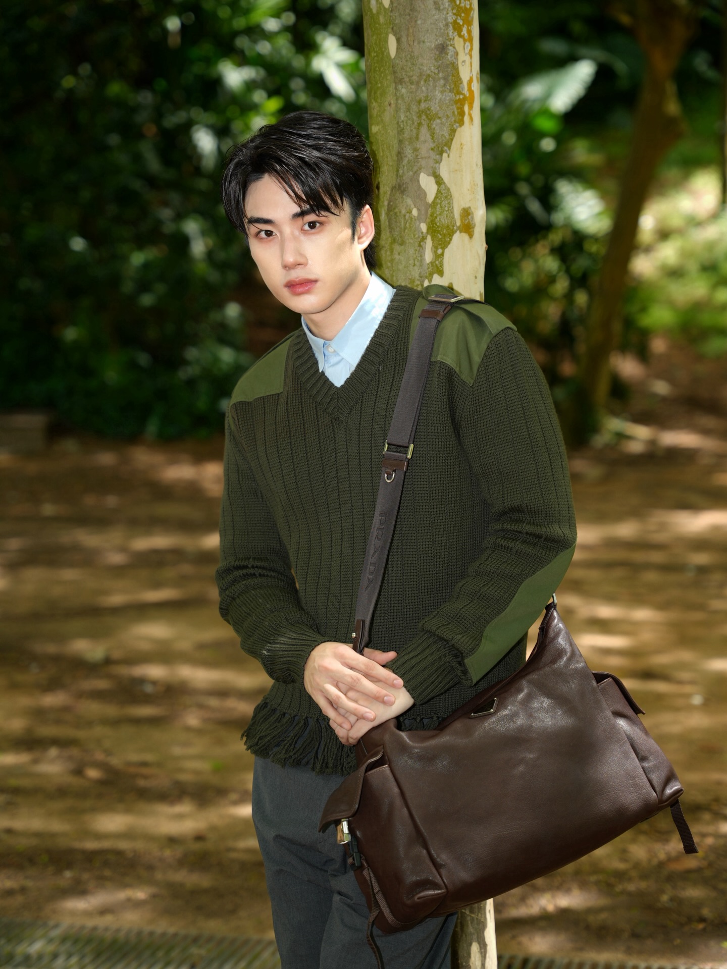 YANG YAN in an olive knit sweater with a leather bag leaning against a tree