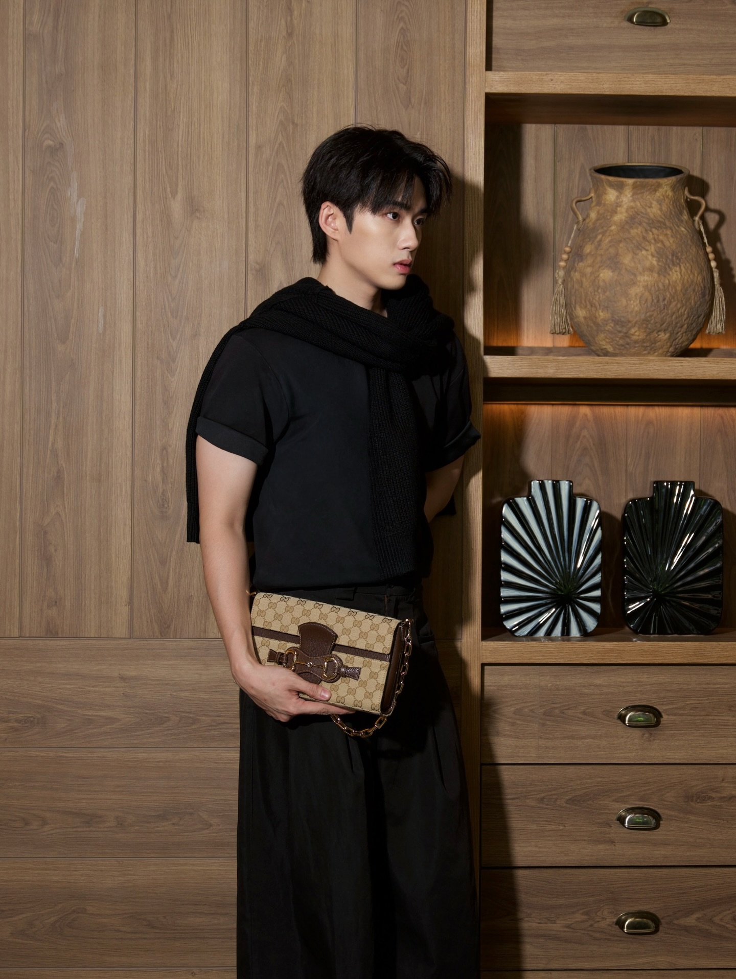 YANG YAN in a black tee with a knit layer over his shoulders holding a monogram clutch against wood panelling