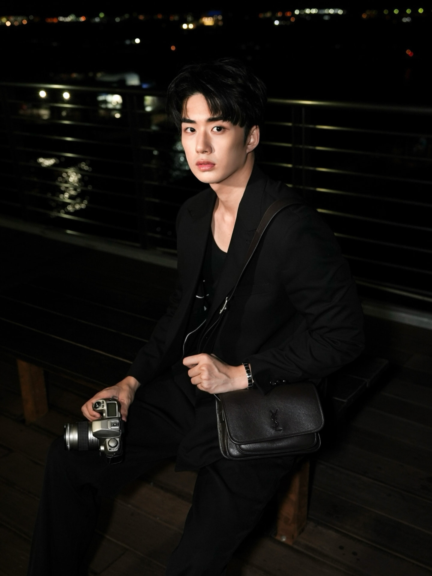 YANG YAN in a black suit holding a film camera on a bench at night